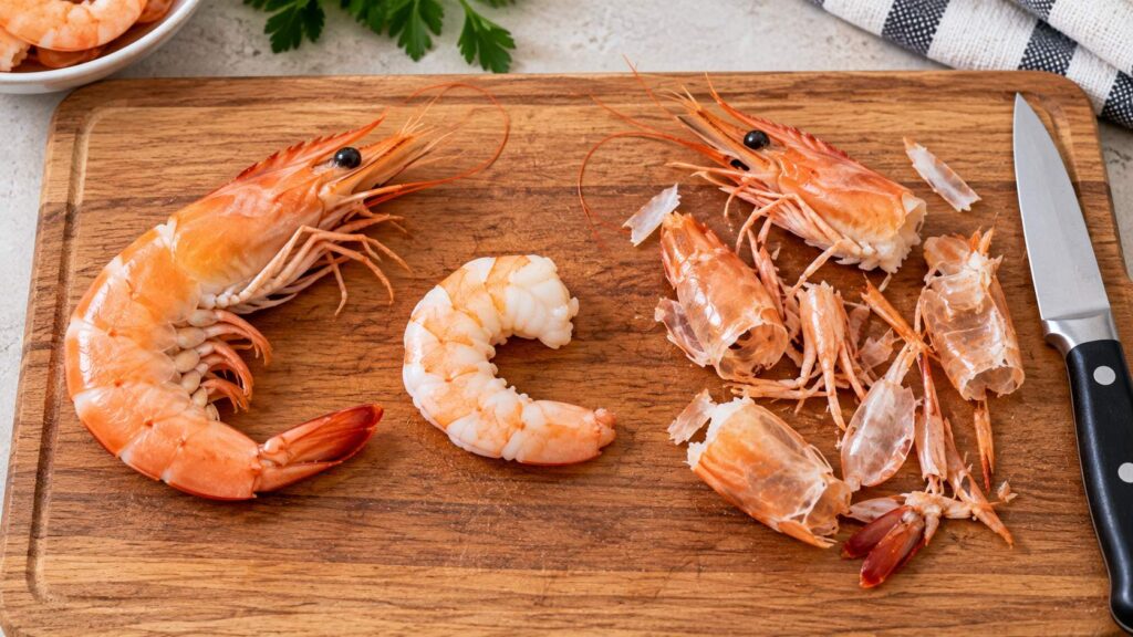 Whole cooked prawn beside peeled prawn meat with the removed shell, head, and tail on a cutting board.